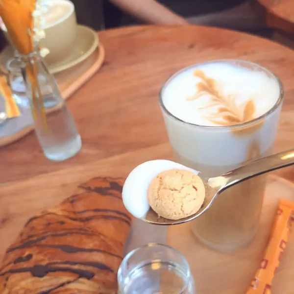 Leckerer Keks mit perfektem Milchschaum auf dem Löffel, im hintergrund ein frisches Schokocroissant und Latte Macchiato
